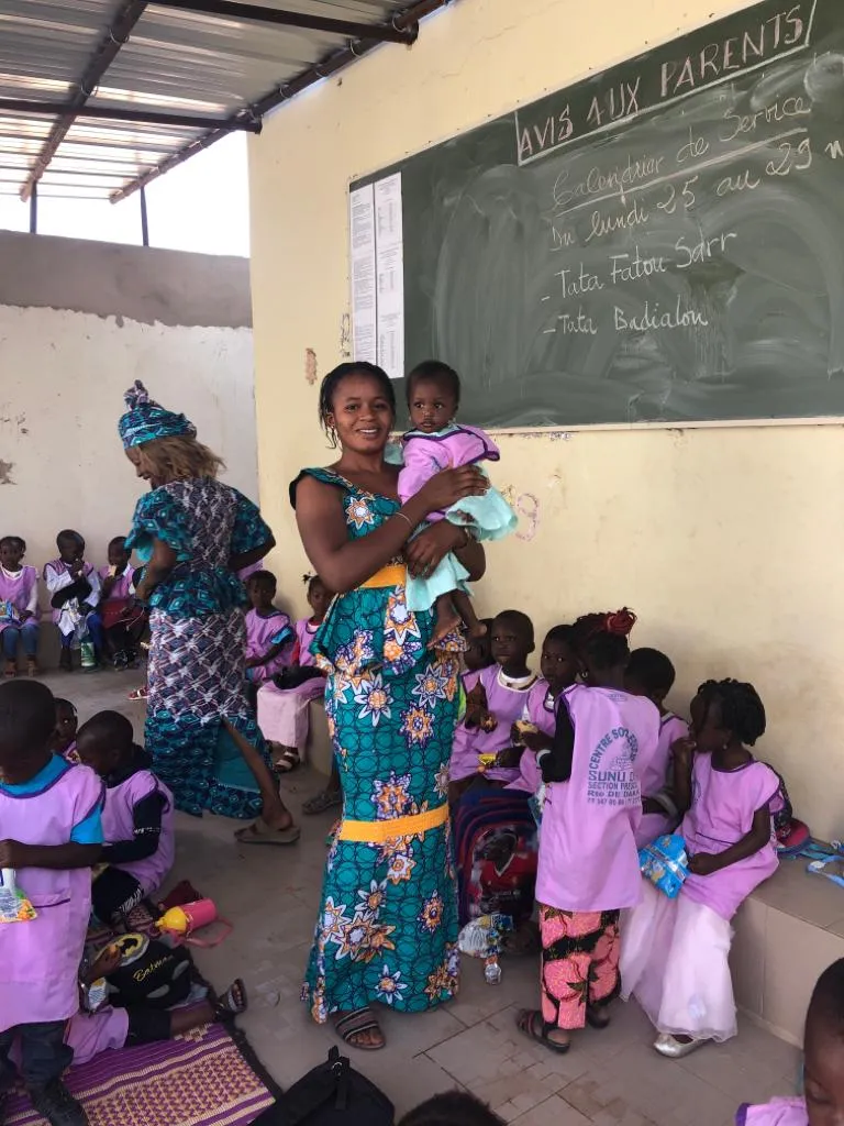 A carer holding a toddler in the kindergarten, children in pink smocks around her and a duty roster on the blackboard