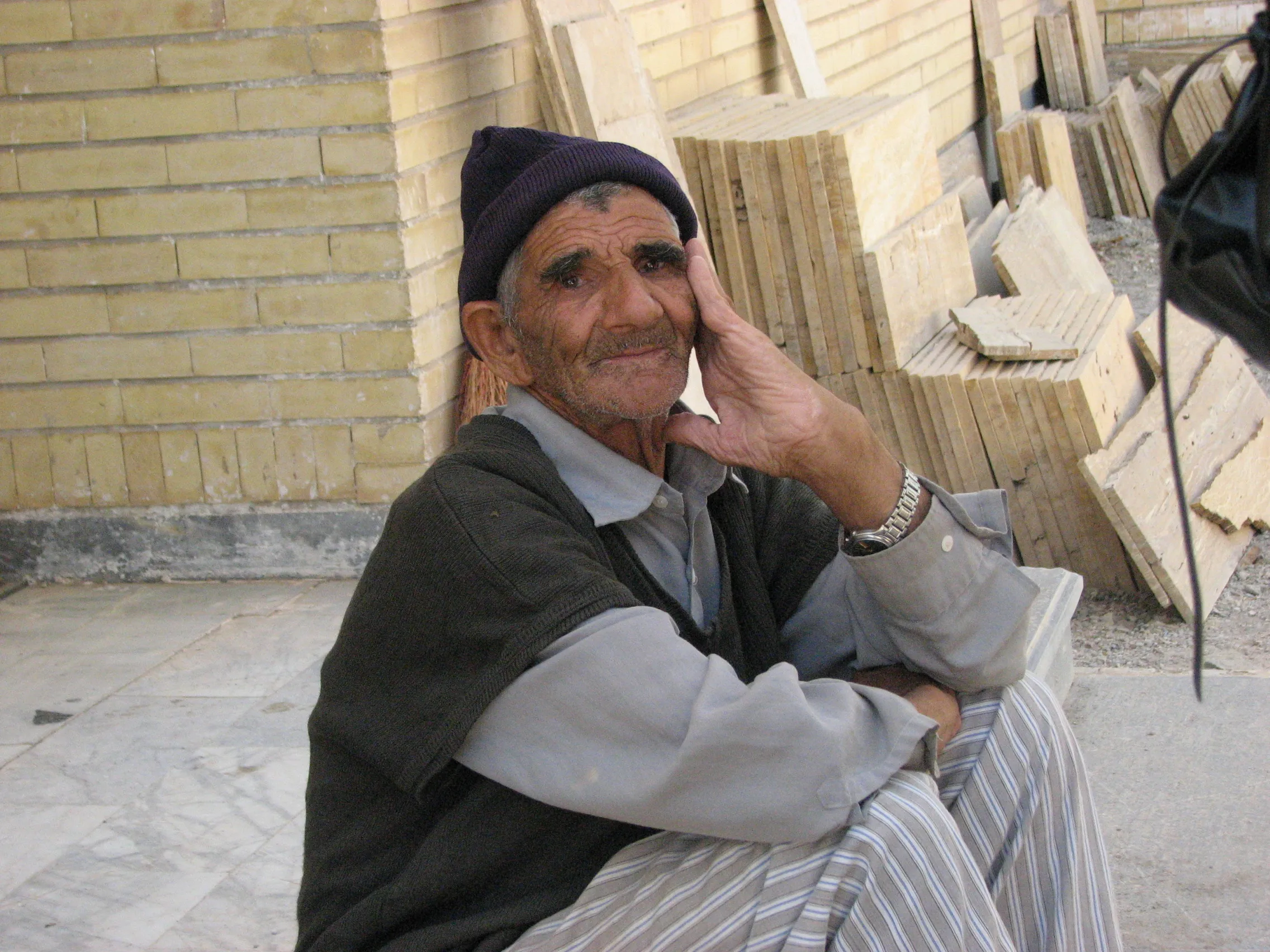 An elderly man resting against a brick wall outside, stacked stone slabs behind him