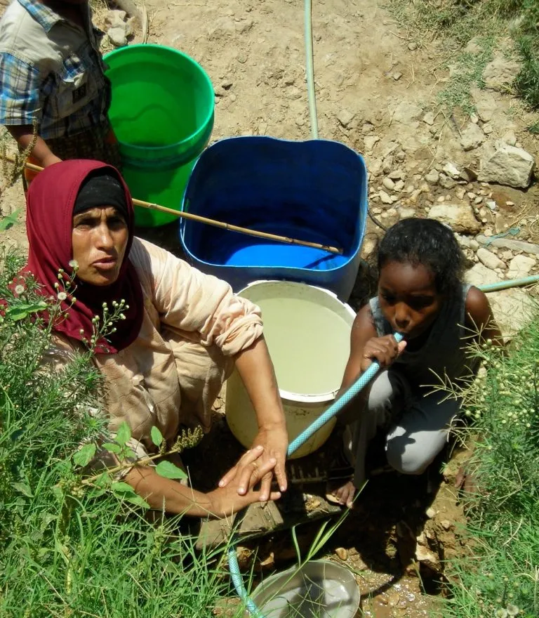 A woman and a girl filling buckets from an open water pipe at the edge of the village, the girl drinking from the hose