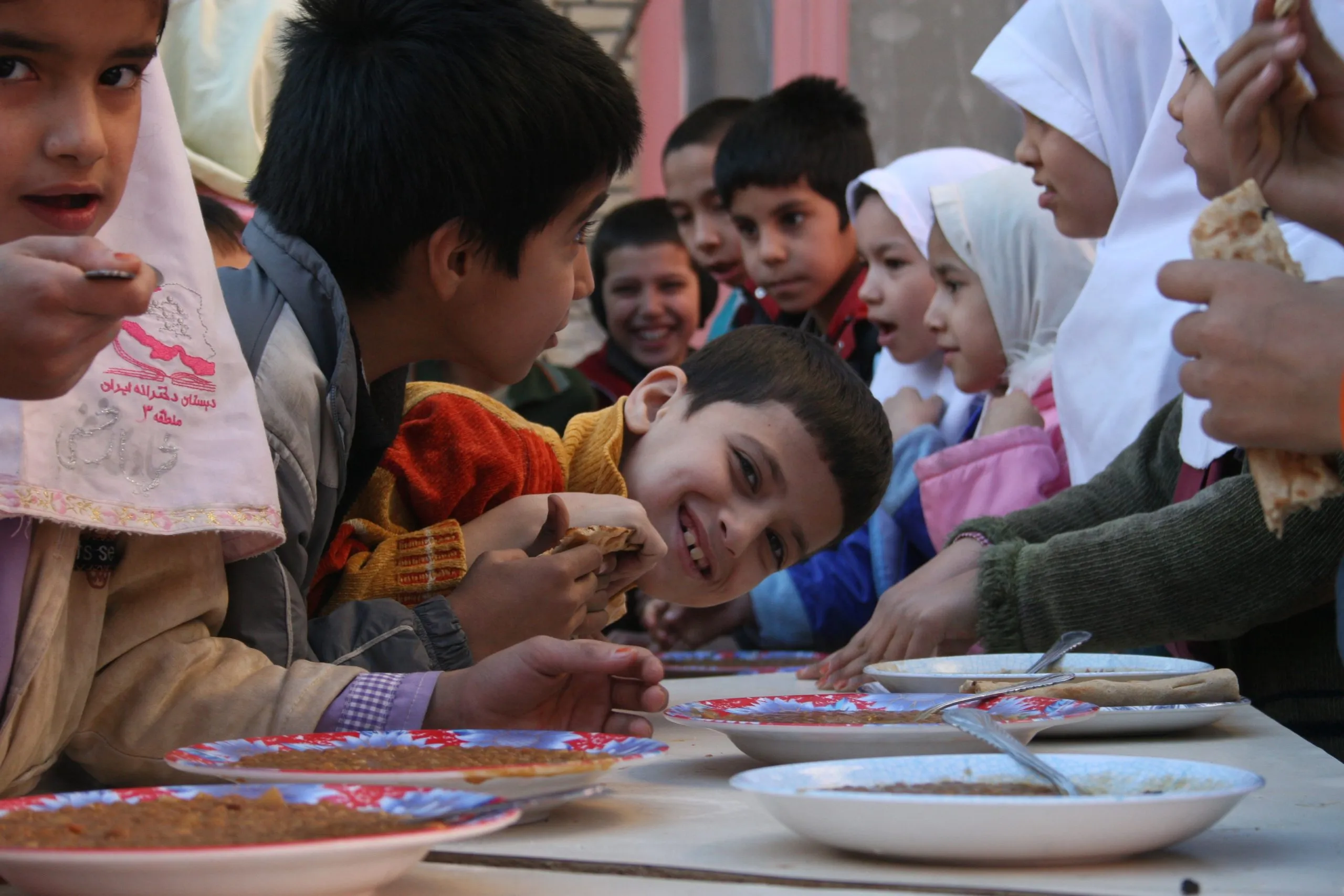 Children crowded along a table eating the hot lunch, bowls of stew and flatbread in front of them