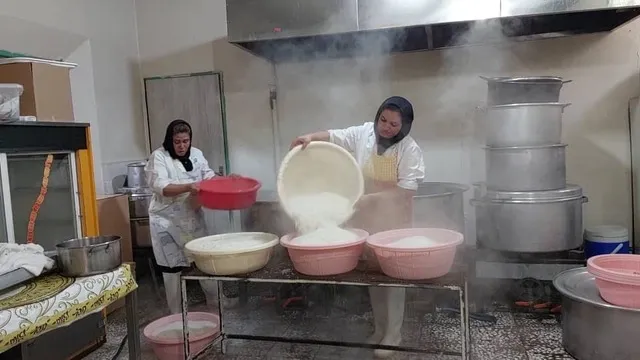Two women in white coats tipping cooked rice from a pot into large bowls in the steamy kitchen