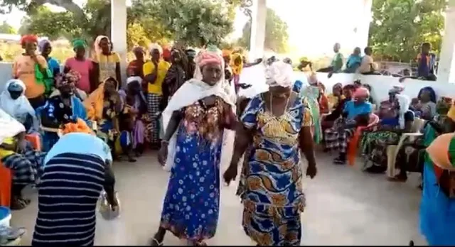 Women dancing at a village gathering, a seated crowd watching