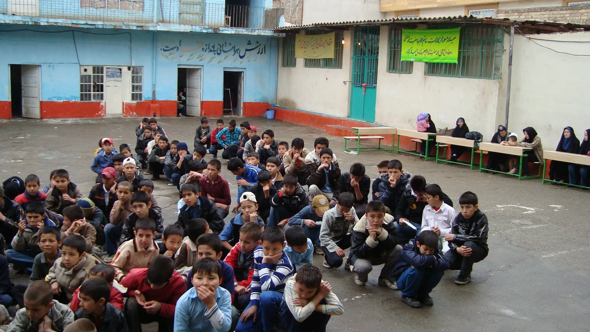 A large group of boys gathered in the school courtyard, with women seated on benches along one side