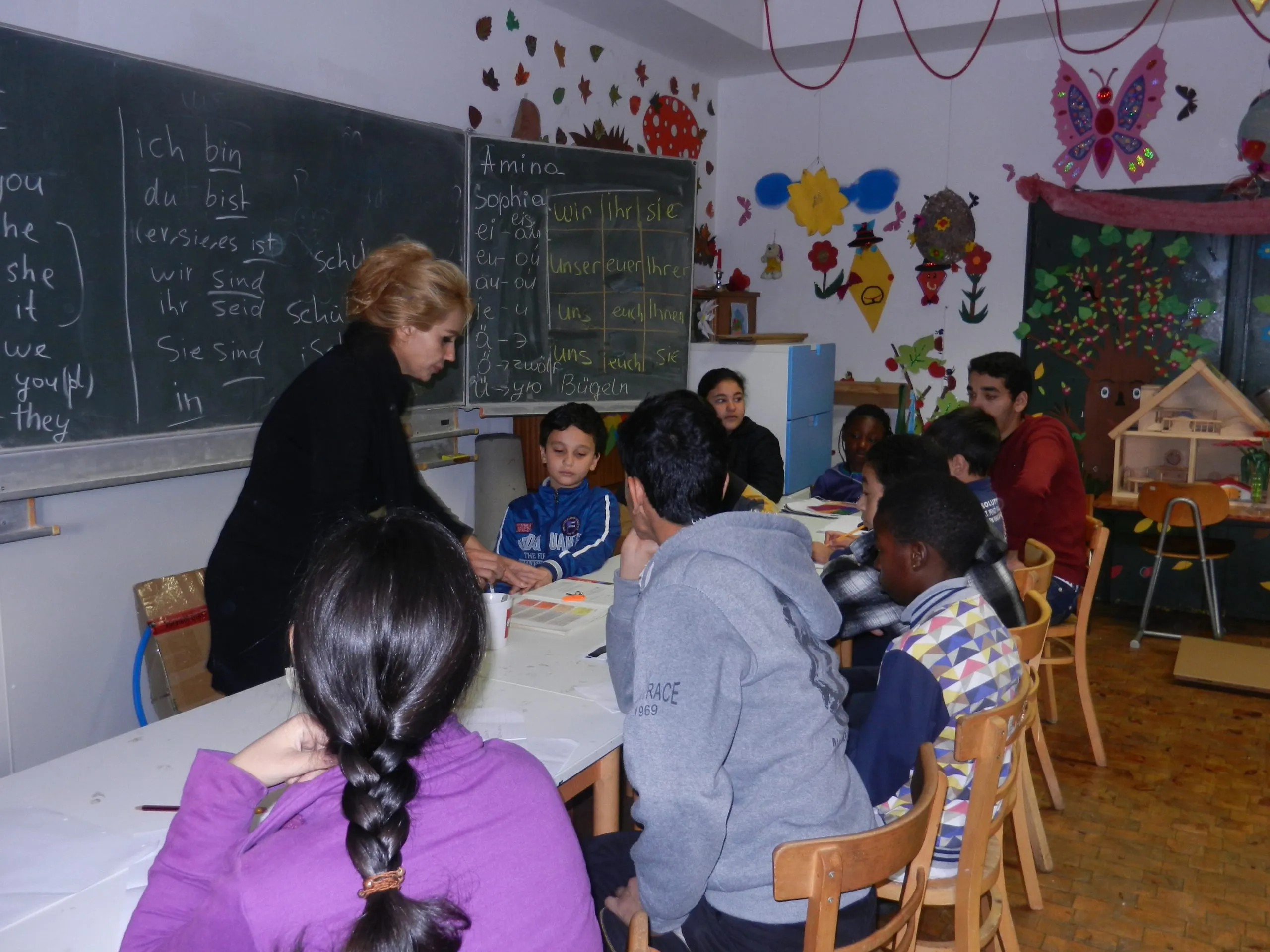 Eine Lehrerin arbeitet mit Kindern und Jugendlichen an einem Tisch vor einer Tafel mit deutschen Pronomen