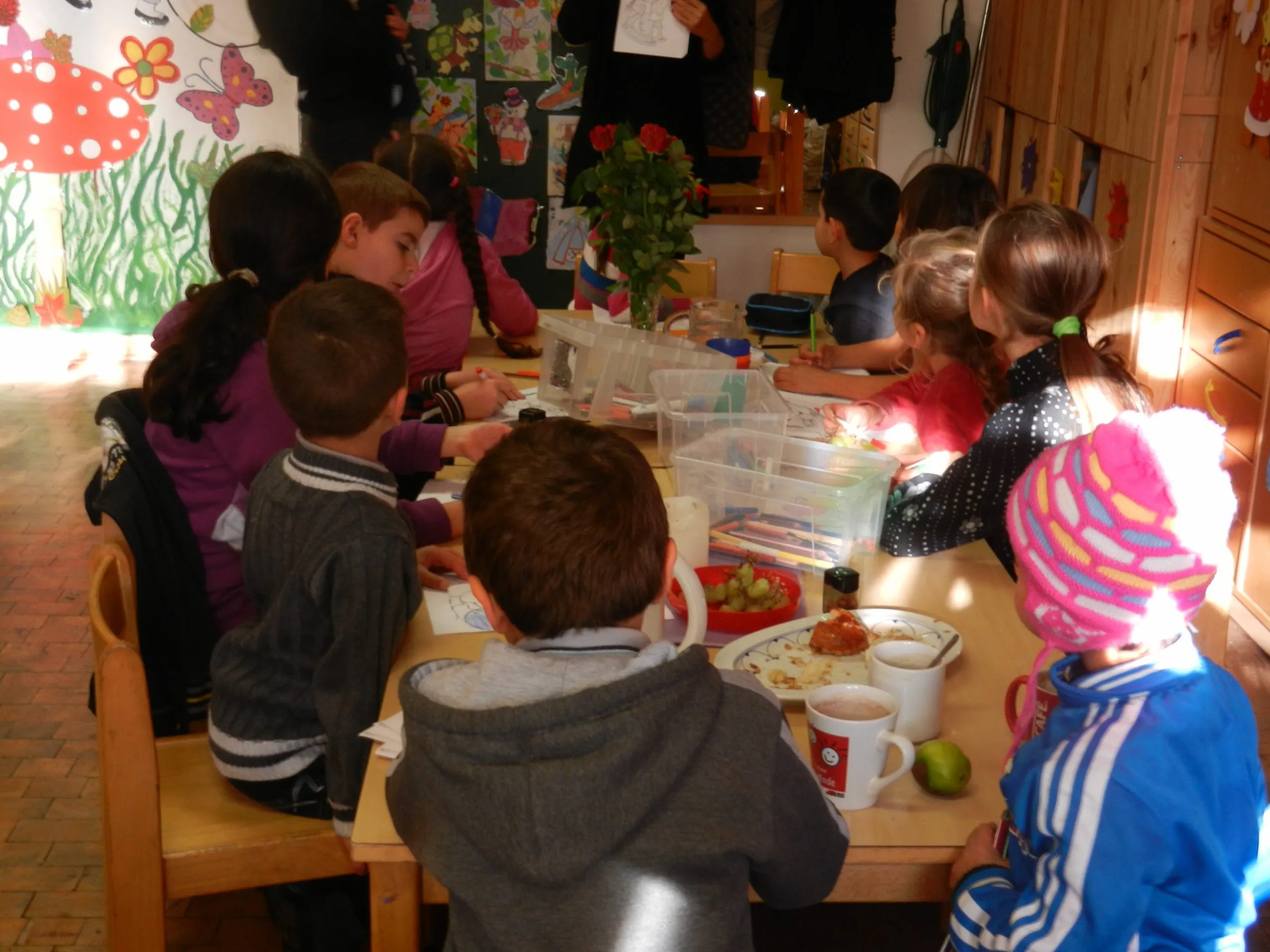 Children drawing and eating a snack around a table in the kindergarten room, a painted mushroom and underwater mural on the wall behind