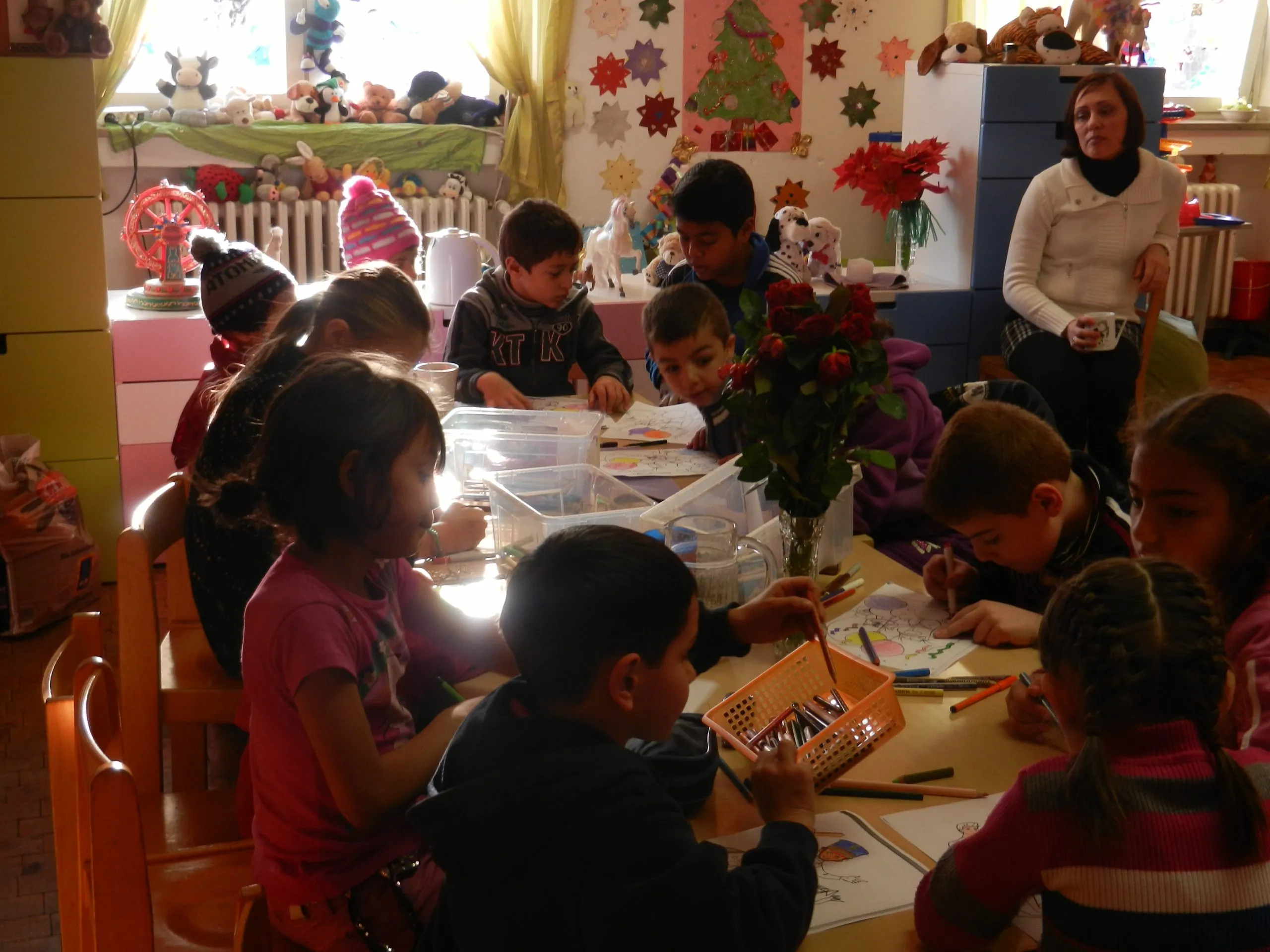 Kinder malen an einem langen Tisch in einem mit Papiersternen und Weihnachtsbaum geschmückten Spielzimmer, im Hintergrund eine Betreuerin