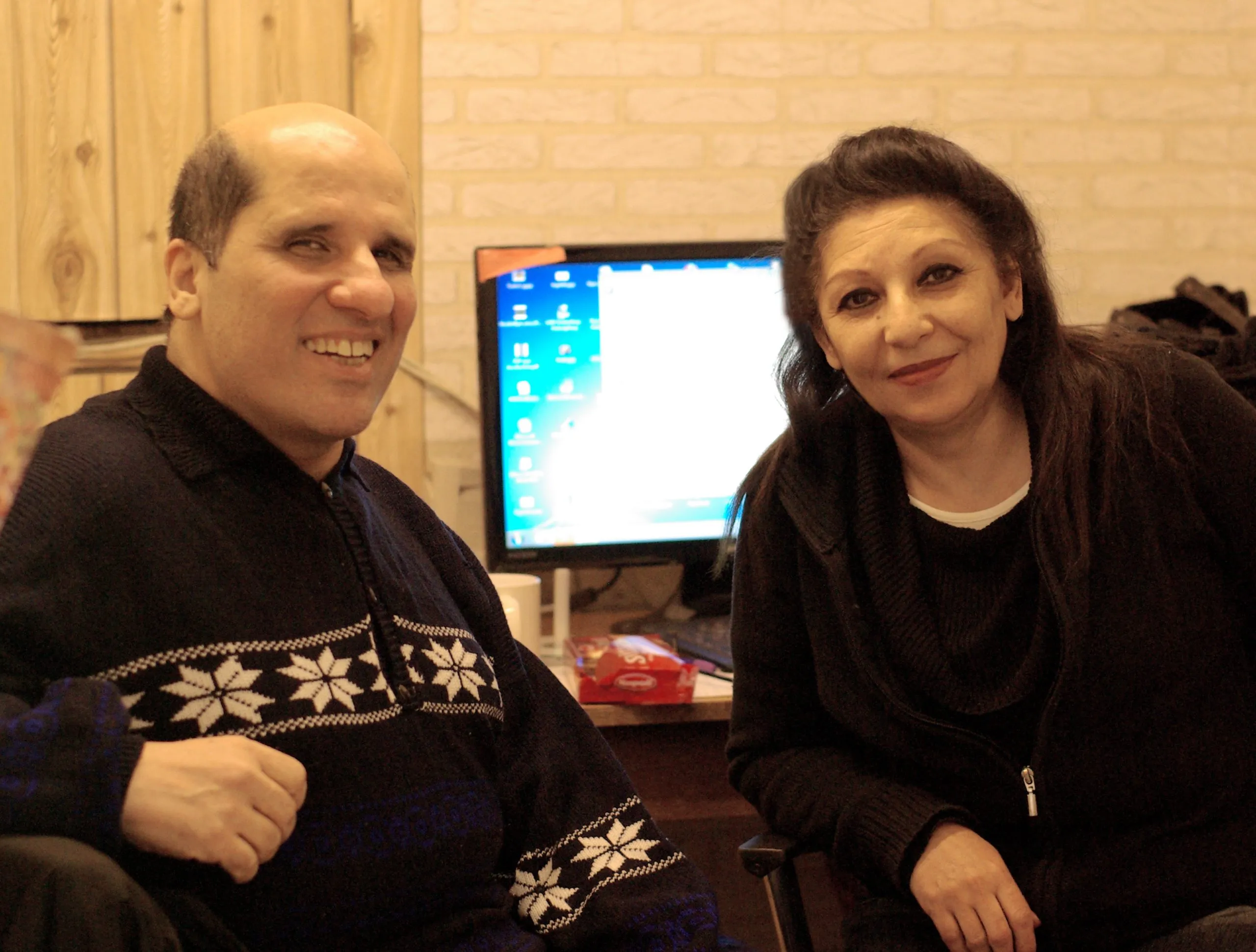 A man and a woman sitting together beside a computer, both looking at the camera