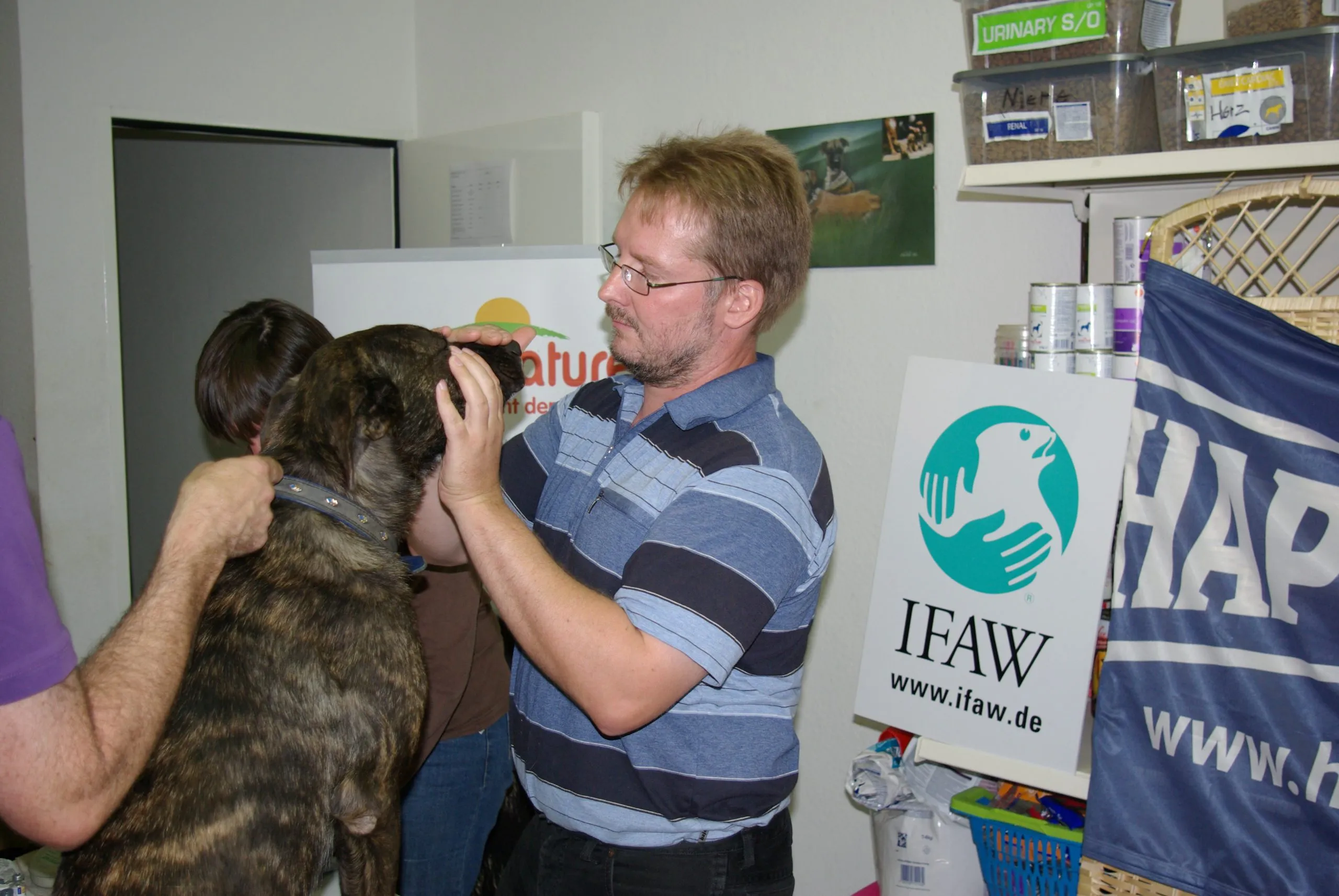 A vet examines a brindle dog held by its owner at a Tiertafel treatment counter, beside shelves of pet food