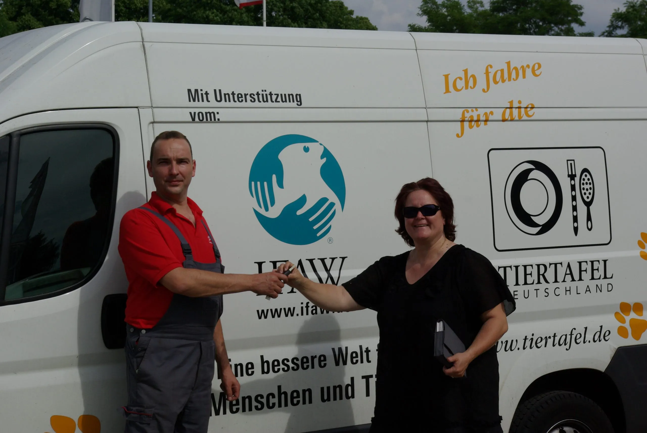 A man hands vehicle keys to a woman in front of a Tiertafel Deutschland van carrying the IFAW logo