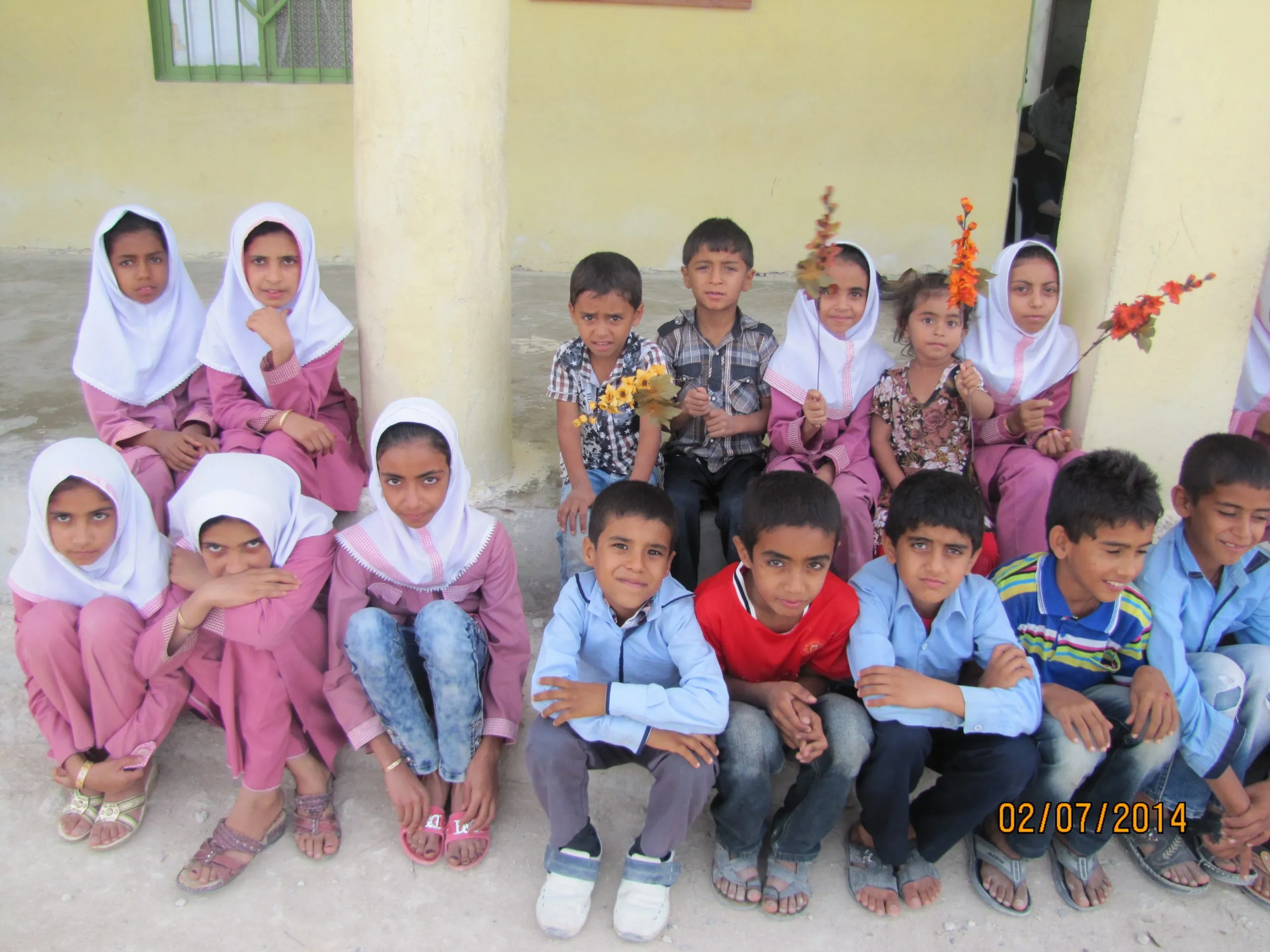 Children sitting on the ground outside the school, several holding flowers