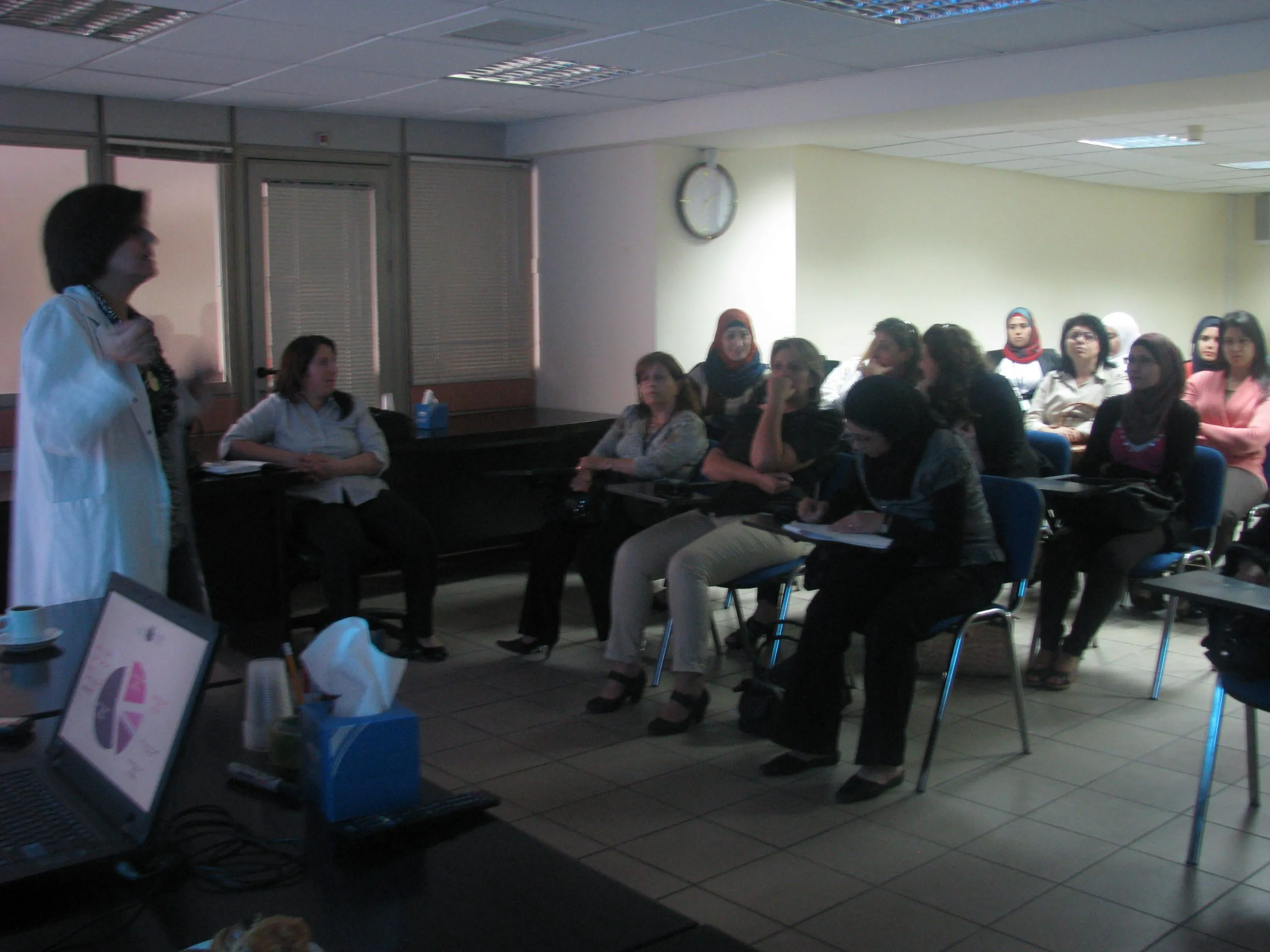 A trainer in a white coat speaking to a seated group of women during a class