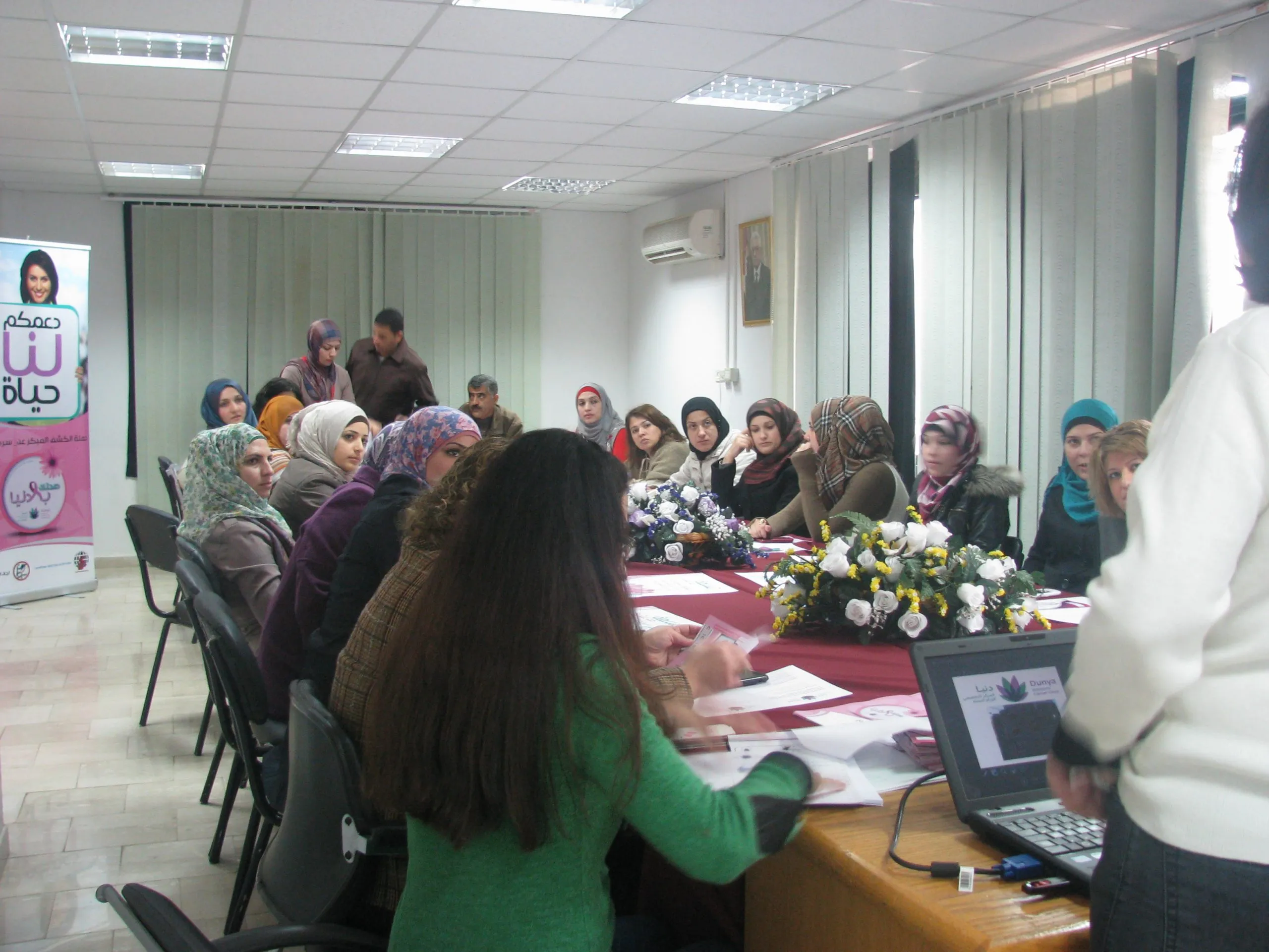 Women seated around a long table at an awareness session, with a Dunya Women's Cancer Center banner at the front