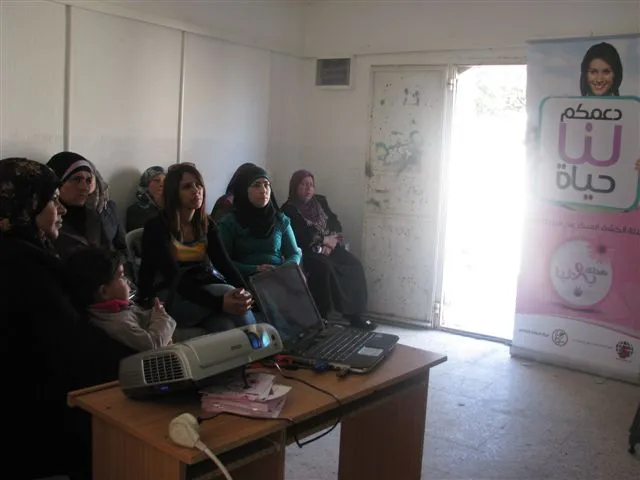 A small group of women watching a projected presentation in a village room, beside a campaign banner