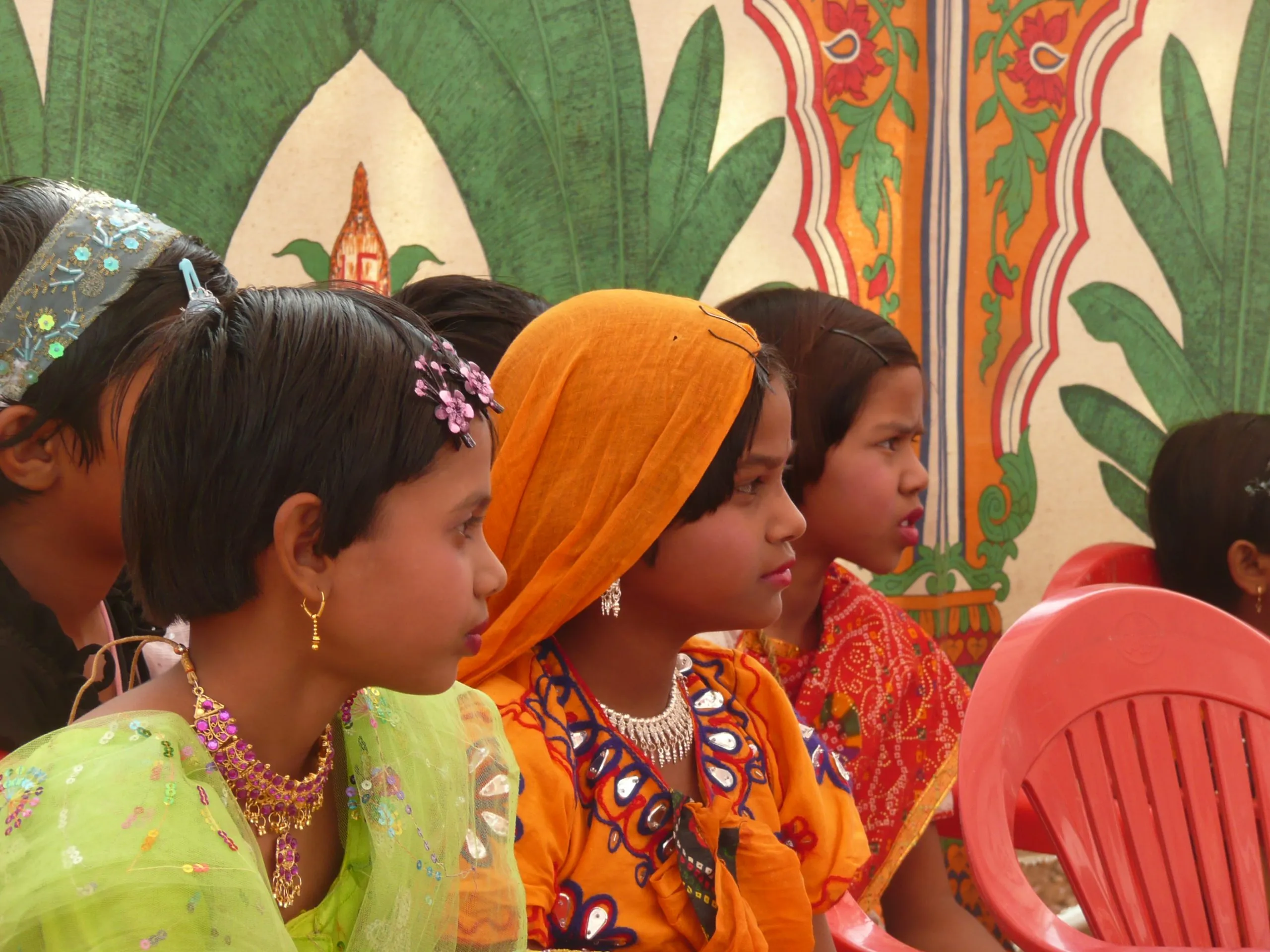 Girls in bright traditional dress and jewellery seated in a row in front of a painted mural