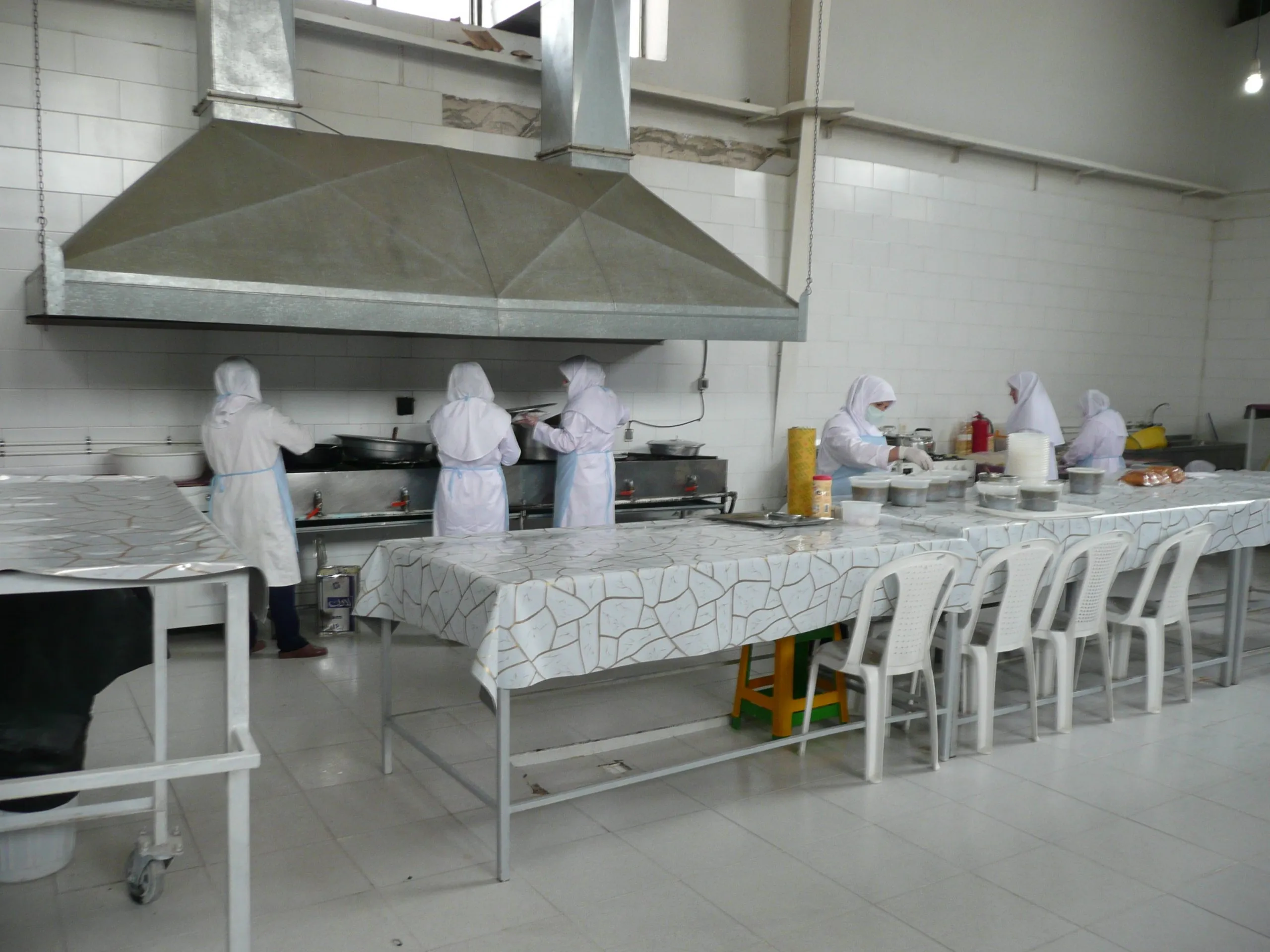 Women in white gowns cooking at a row of gas burners and portioning meals into containers