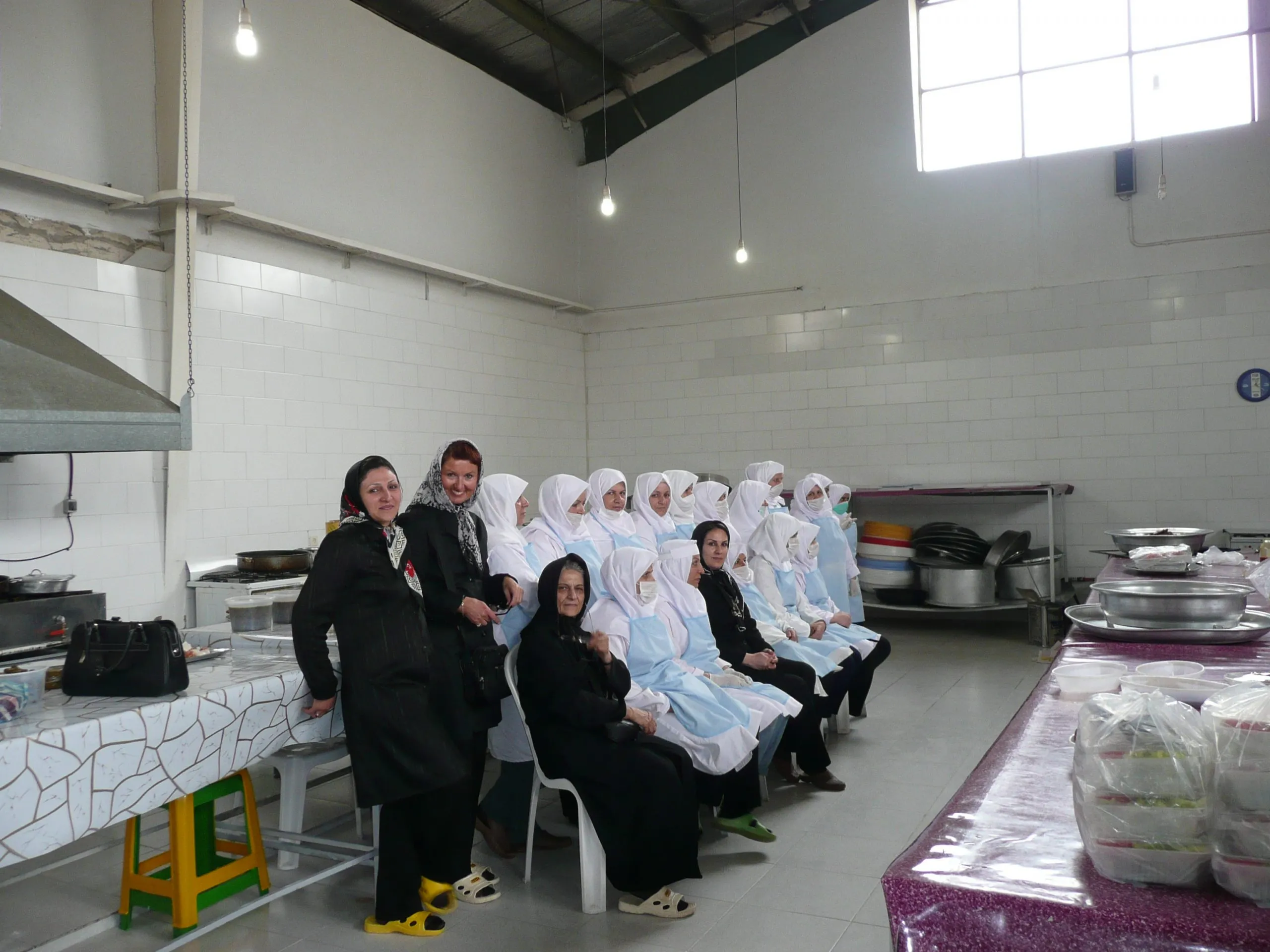 Women in white hygiene gowns and headscarves seated in the project kitchen, with two visitors standing beside them