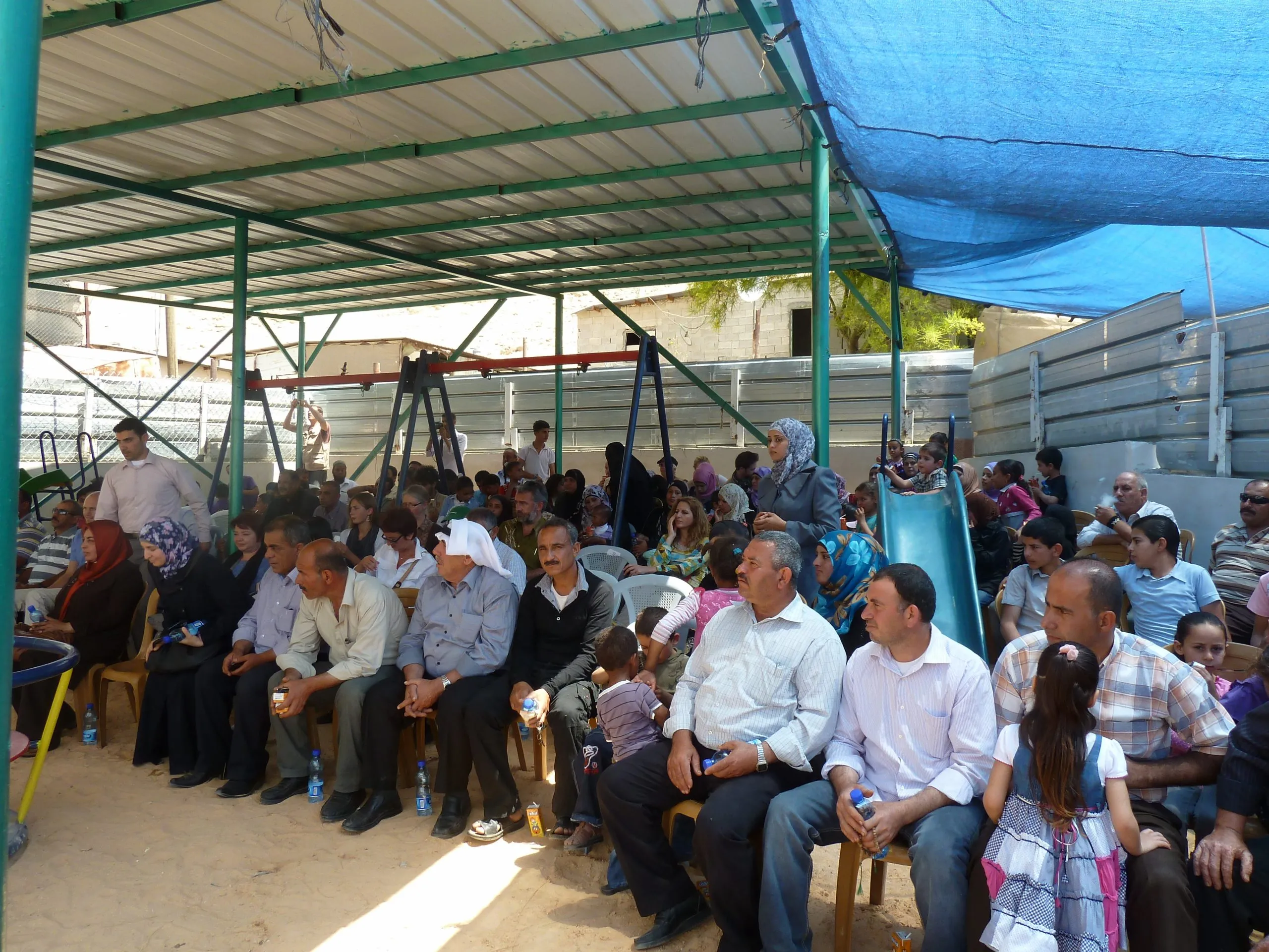 Villagers seated under a shaded canopy at the kindergarten opening, play equipment behind them