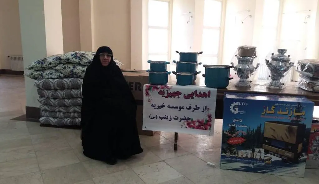 A woman seated beside donated household goods — bedding, pans, samovars and a gas cooker — with a sign naming the Zainab charity