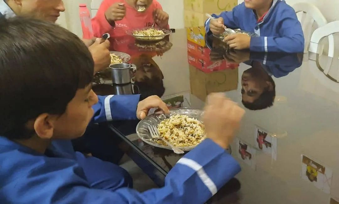 Kinder in blauen Schulkitteln essen an einem Glastisch einen Teller Reis