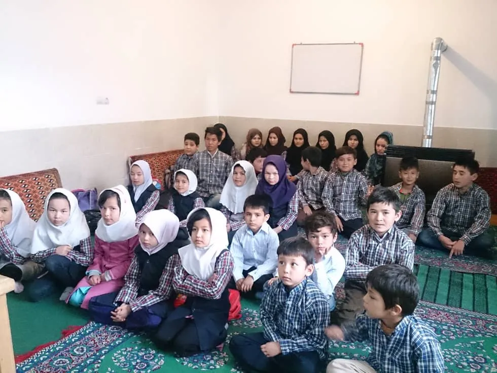 Children in checked school shirts and white headscarves sitting together on the classroom floor