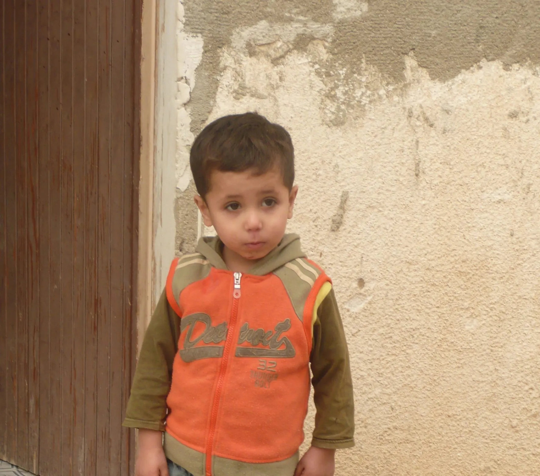 A small boy in an orange hooded top standing in a doorway against a weathered wall