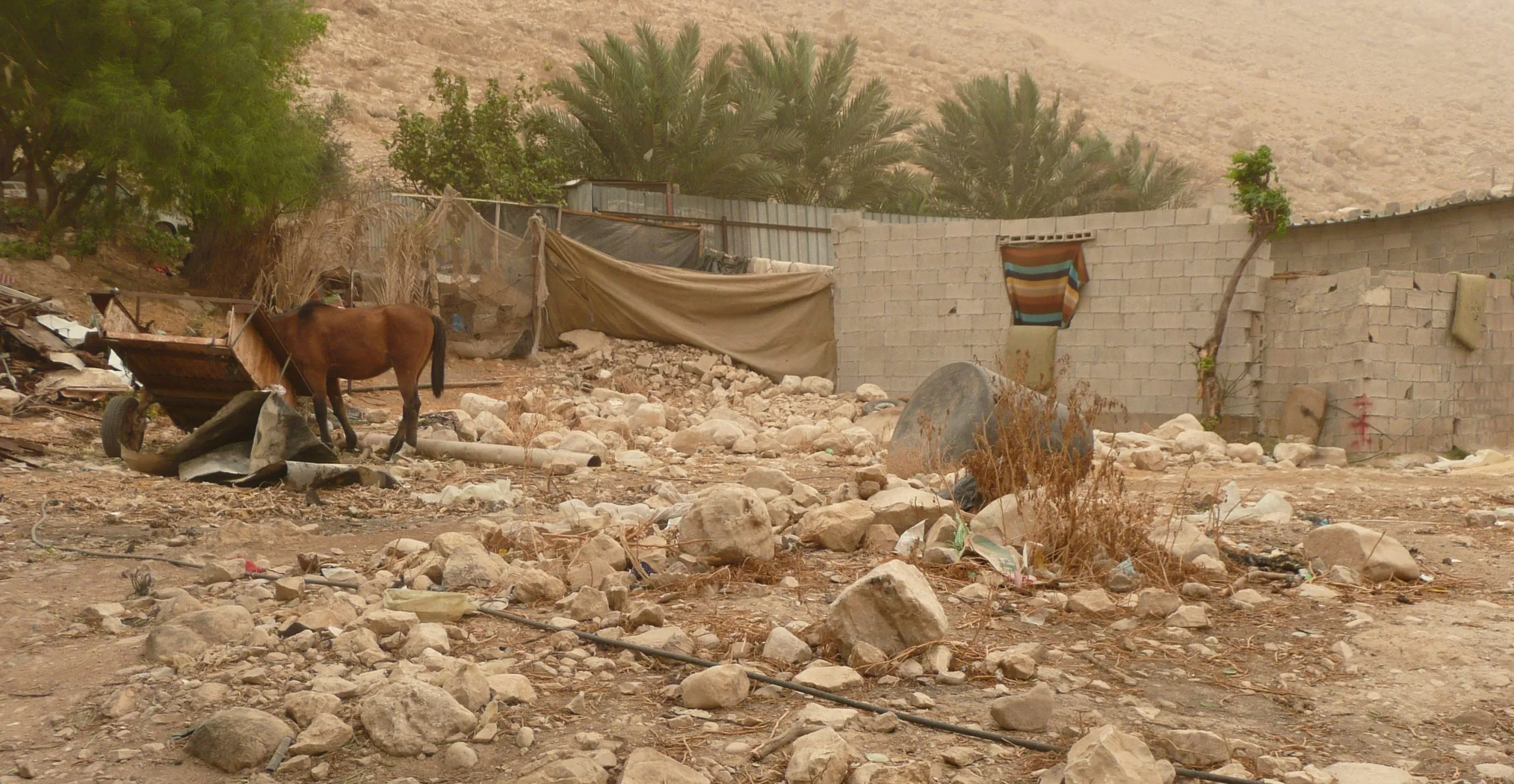 A dusty village yard with a horse and cart among rubble, desert hills rising behind